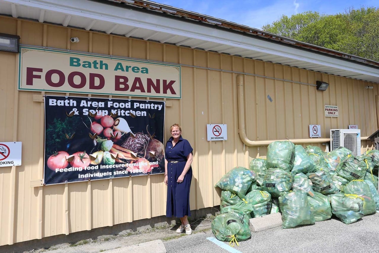Person standing outside Bath Area Food Bank with bags of supplies, next to a sign for food pantry and soup kitchen.