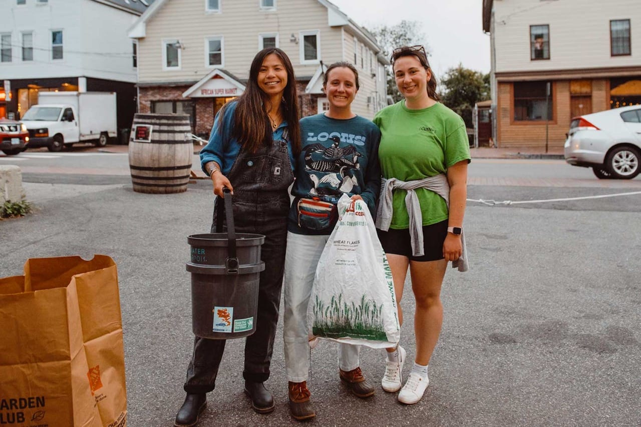 Three people smiling while holding a bucket and a bag, standing outdoors in front of buildings and parked cars.