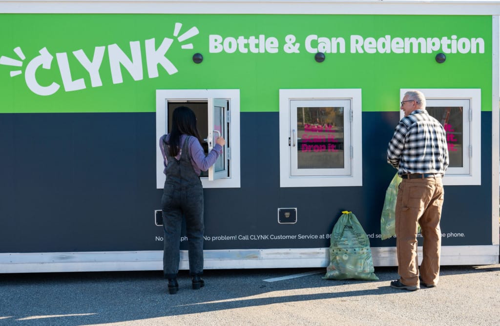 Two people returning bottles at a CLYNK redemption center with a green and black exterior and service windows.